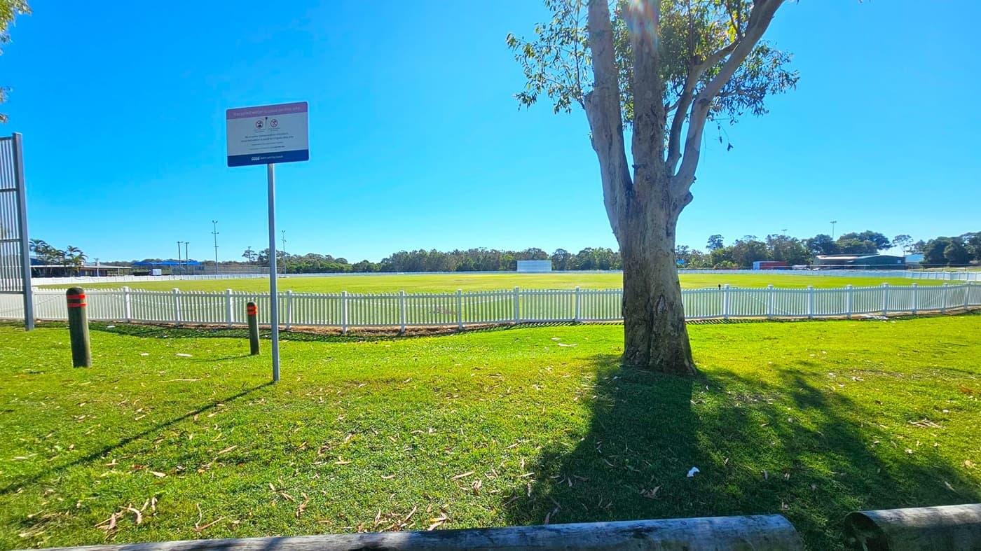 A grassy field with a white picket fence in the background. A large tree stands on the right, casting a shadow on the grass. A signpost is visible to the left, near two bollards. In the distance, there are trees and a few buildings. The sky is clear and blue.