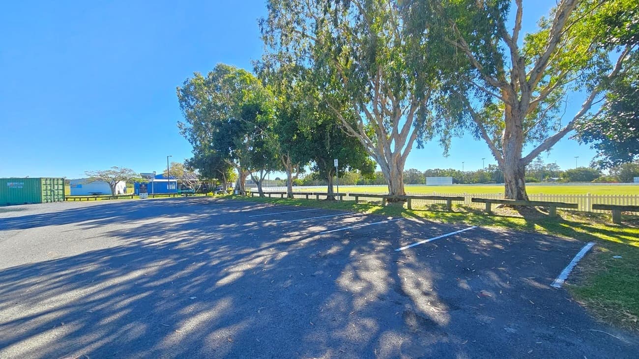 A car park with marked spaces on an asphalt surface, partially shaded by large trees on the right. To the left, there are green shipping containers and a small blue building. In the background, a grassy field is enclosed by a white picket fence. Shadows of the trees are cast on the ground, and a few benches are placed along the fence line.