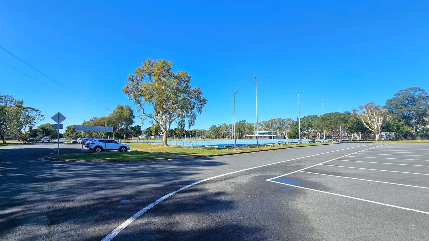 An empty car park with marked spaces on the right. A few cars are parked on the left near a road, with a white car visible. Several trees are scattered throughout the area, providing some shade. In the background, there is a fenced sports field with tall floodlights and a building. The ground is paved, and the sky is clear.