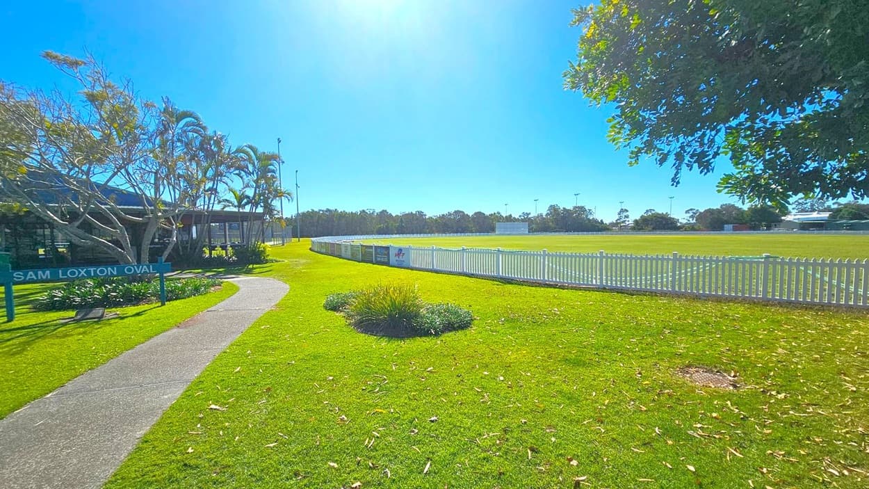A grassy area with a curved concrete path leading to a building on the left, surrounded by palm trees and other greenery. A sign reads "Sam Loxton Oval." On the right, a white picket fence encloses a large, well-maintained sports field. Trees and shrubs are visible in the background, with a few tall light poles around the field. The foreground has scattered leaves on the grass, and a tree with dense foliage partially shades the scene.
