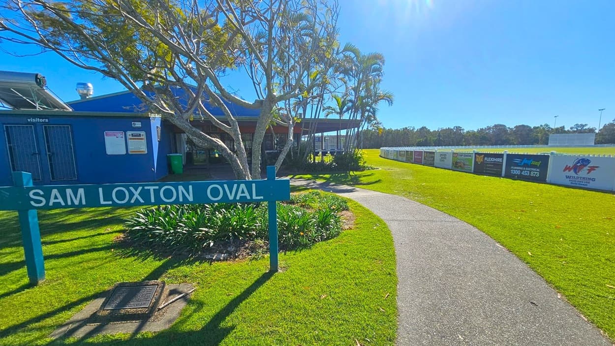 A sign reading "Sam Loxton Oval" is in the foreground on the left, surrounded by green plants and grass. Behind it, a blue building with a "visitors" sign is partially visible, with a tree providing shade. A pathway curves to the right, leading towards a grassy field bordered by a white fence with various advertisements. Tall trees and a clear sky are in the background.