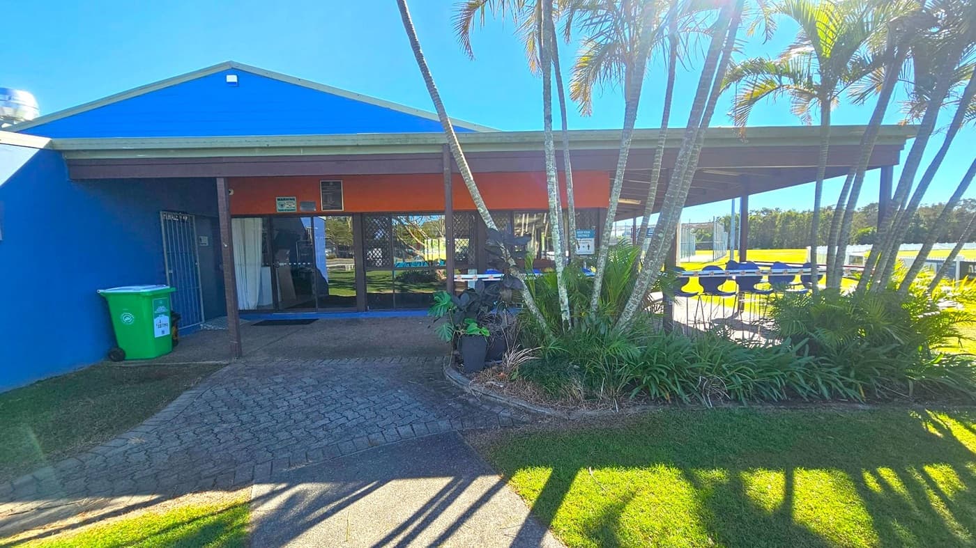 A building with a blue exterior and a sloped roof. The entrance has glass doors and a small overhang. A green recycling bin is on the left near a gated door. On the right, there is a covered outdoor seating area with blue tables and chairs. Several palm trees and plants are in the foreground, casting shadows on the grass and paved path. In the background, there is a grassy field and a line of trees.