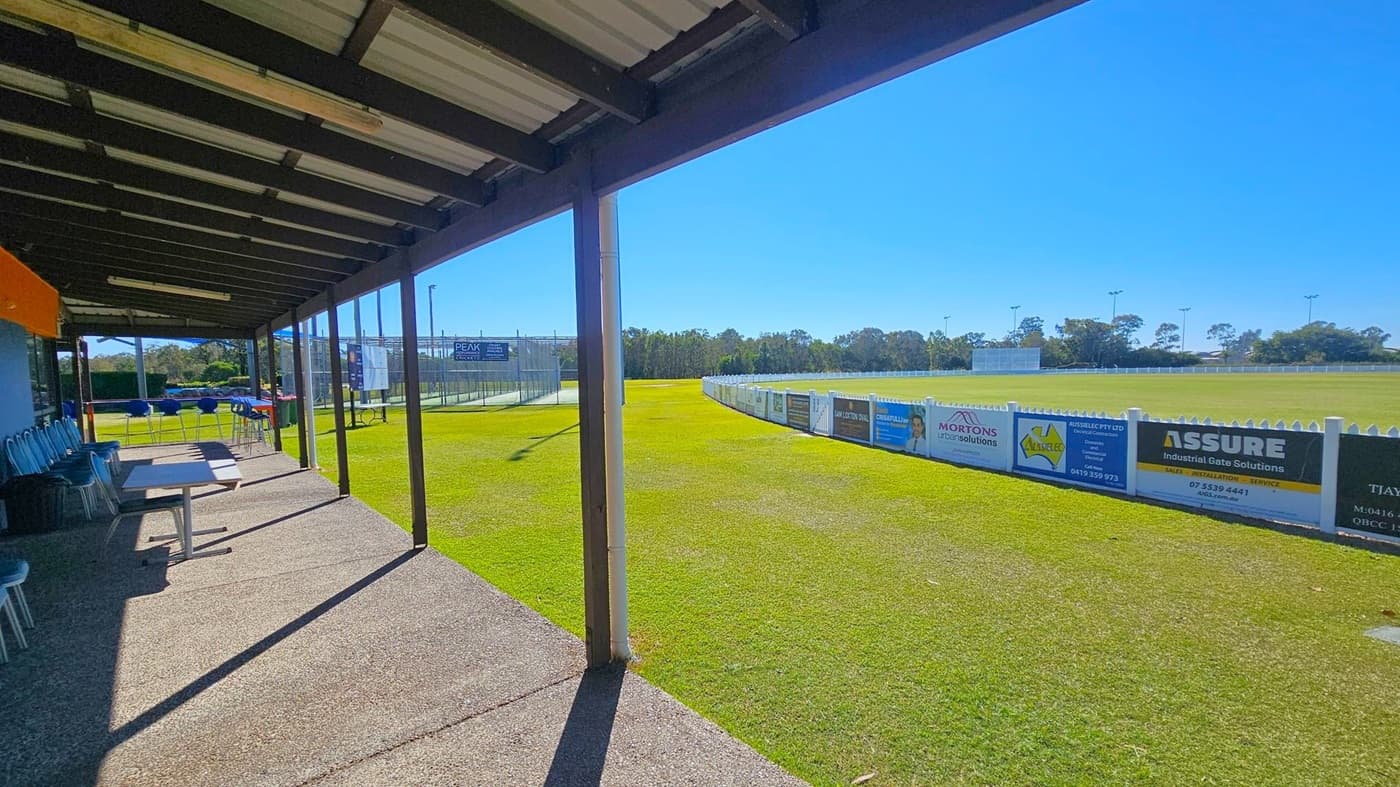 A covered outdoor area with a metal roof and concrete floor on the left, featuring several chairs and tables. Beyond the covered area, a grassy sports field extends to the right, bordered by a white fence with advertising banners. In the background, there are trees and a cricket sight screen. The sky is clear and blue.
