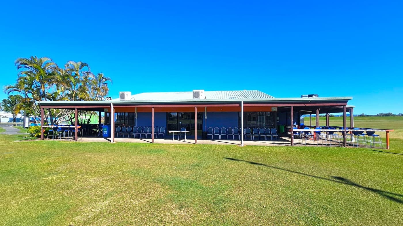 A single-storey building with a metal roof and a covered veranda runs across the front. On the left, there are tall palm trees next to the building. The veranda has a row of chairs lined up against the wall, and tables with chairs are set up on both the left and right sides. The ground is covered with grass, and the sky is clear. The building has air conditioning units on the roof.