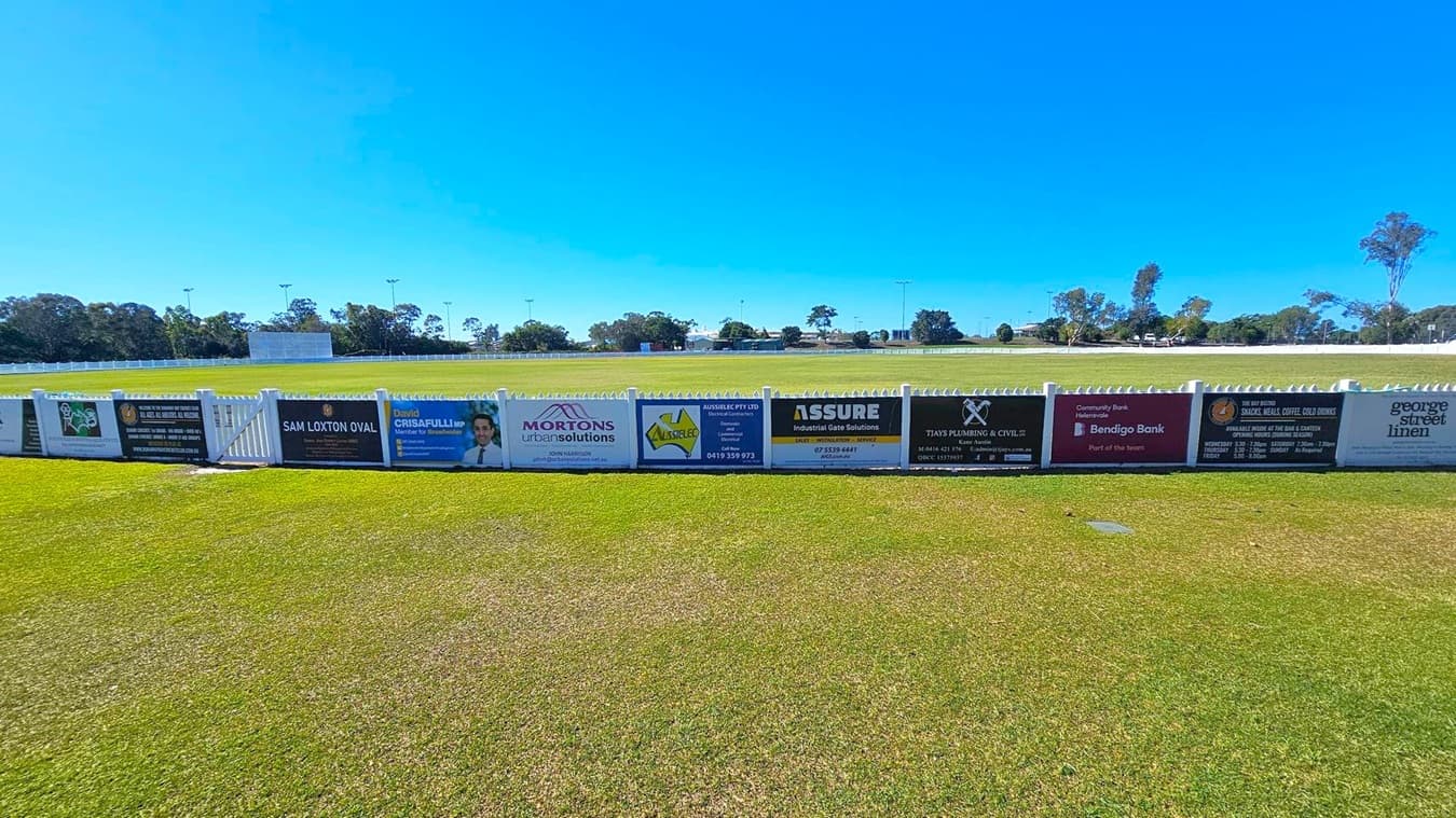 A grassy sports field with a white picket fence in the foreground, displaying various advertising signs. The signs include names and logos of businesses such as Mortons Urban Solutions, Bendigo Bank, and others. In the background, there are trees and a scoreboard. The field is expansive, with light poles visible in the distance. The sky is clear and blue.
