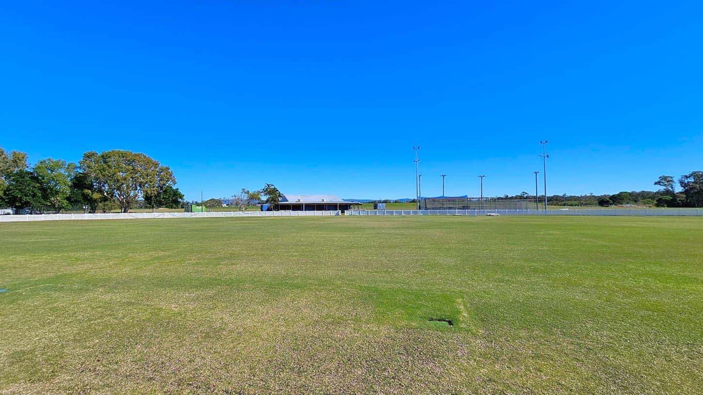 A wide, open grassy field with a well-maintained lawn. In the background, there is a low building with a light-coloured roof, surrounded by a white fence. To the left, several large trees provide some greenery. On the right, tall light poles are visible, likely for illuminating the area. The field is expansive, with a few patches of different shades of green and brown on the grass.
