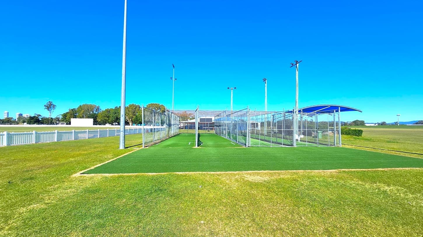 Cricket practice nets with green artificial turf in the centre, surrounded by metal fencing. Tall floodlights are positioned around the area. To the left, a white picket fence borders a grassy field with trees and distant buildings. On the right, a blue curved shelter is visible, with more open grassy space extending into the distance. The ground is a mix of green and brown grass.