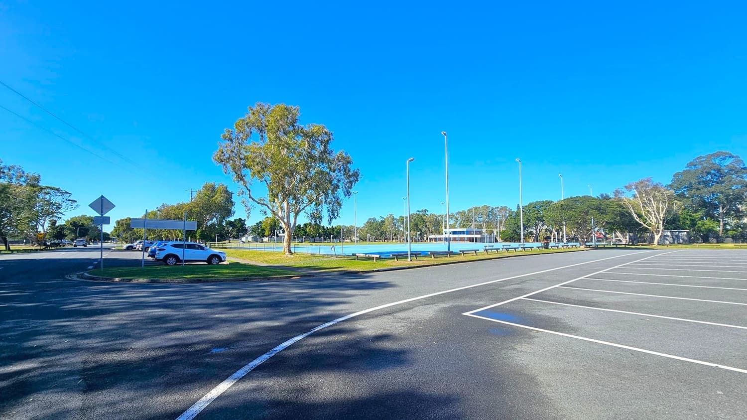 A large, empty car park with white line markings on the right. In the middle, a few cars are parked near a grassy area with several tall trees. A road curves around the left side, with a few road signs visible. In the background, there is a fenced sports field with tall floodlights and a building surrounded by more trees. The sky is clear and blue.