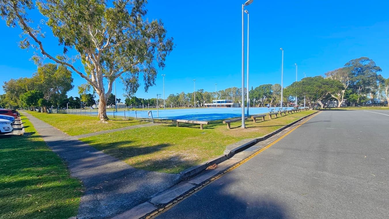 A paved road runs along the right side, bordered by a grassy area with a row of wooden benches. On the left, several parked cars line a path shaded by tall trees. In the centre, a fenced sports court with a blue surface is visible, surrounded by tall floodlights. Trees and a building are in the background, with clear blue skies above.