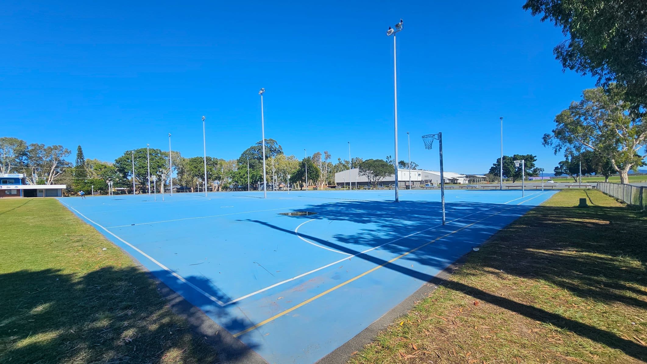 A blue outdoor netball court with multiple tall light poles. On the left, there's a grassy area leading to a building with a white roof. Trees with green foliage are scattered in the background. On the right, a fence runs alongside more grass and trees. Shadows from the trees and poles stretch across the court. The court has white and yellow lines, and a netball hoop is visible.