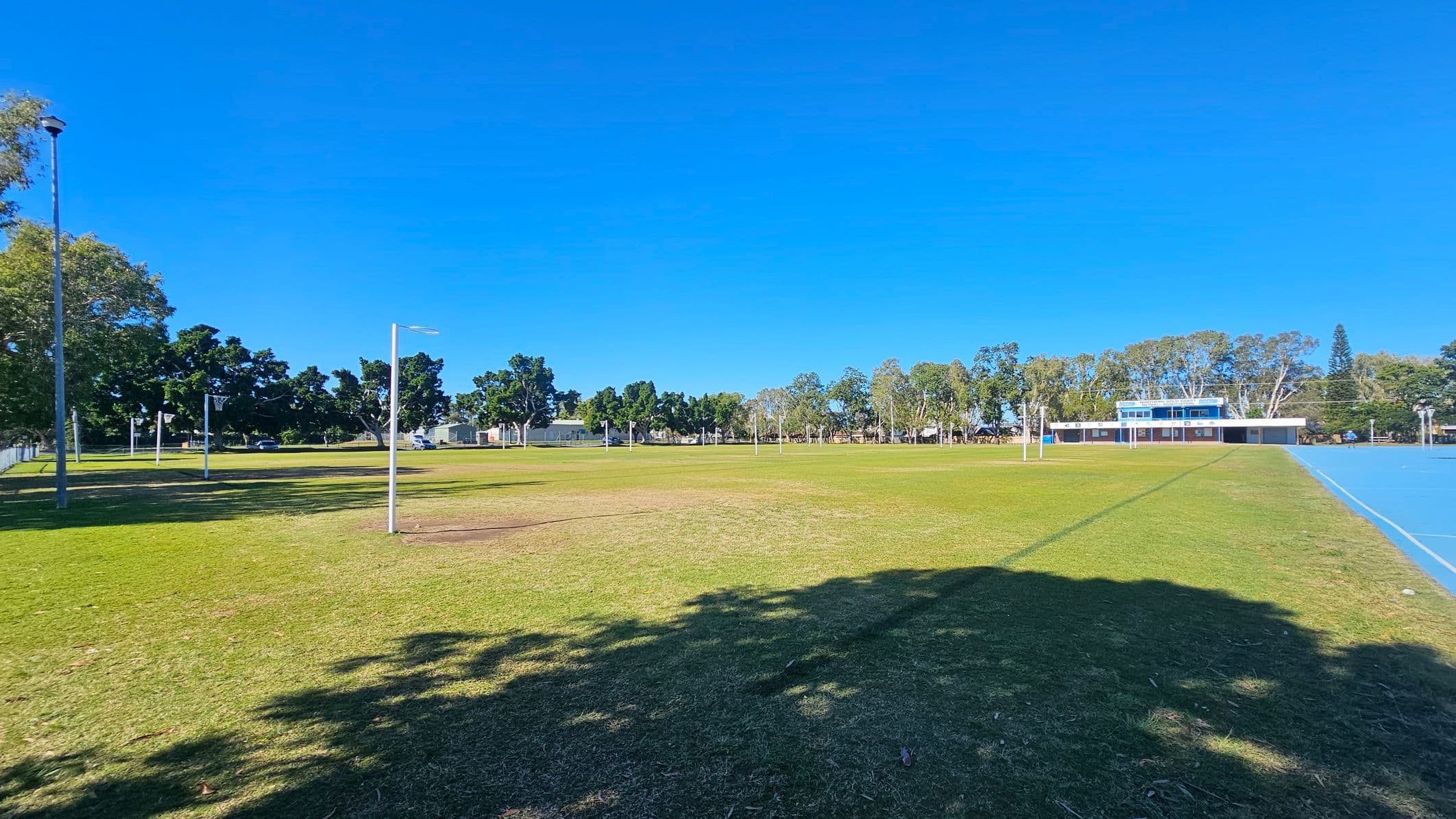 A grassy field with scattered trees and several tall light poles. On the right, a blue sports court runs parallel to the field. In the background, a building with a blue roof is partially visible, surrounded by more trees. Shadows from the trees are cast on the grass in the foreground.