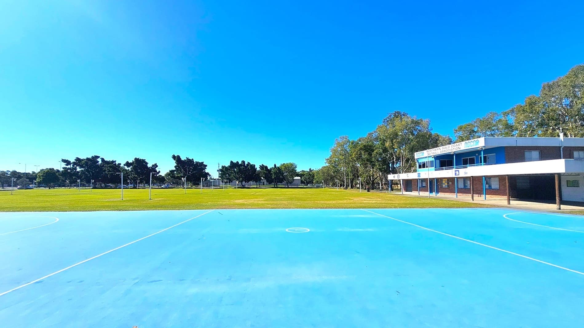 A blue netball court occupies the foreground, with white lines marking the playing area. In the background, a grassy field is bordered by trees. On the right, a building with signage for the Northern Gold Coast Netball Association is visible, featuring a white and blue facade. The building has multiple windows and a covered walkway supported by columns.