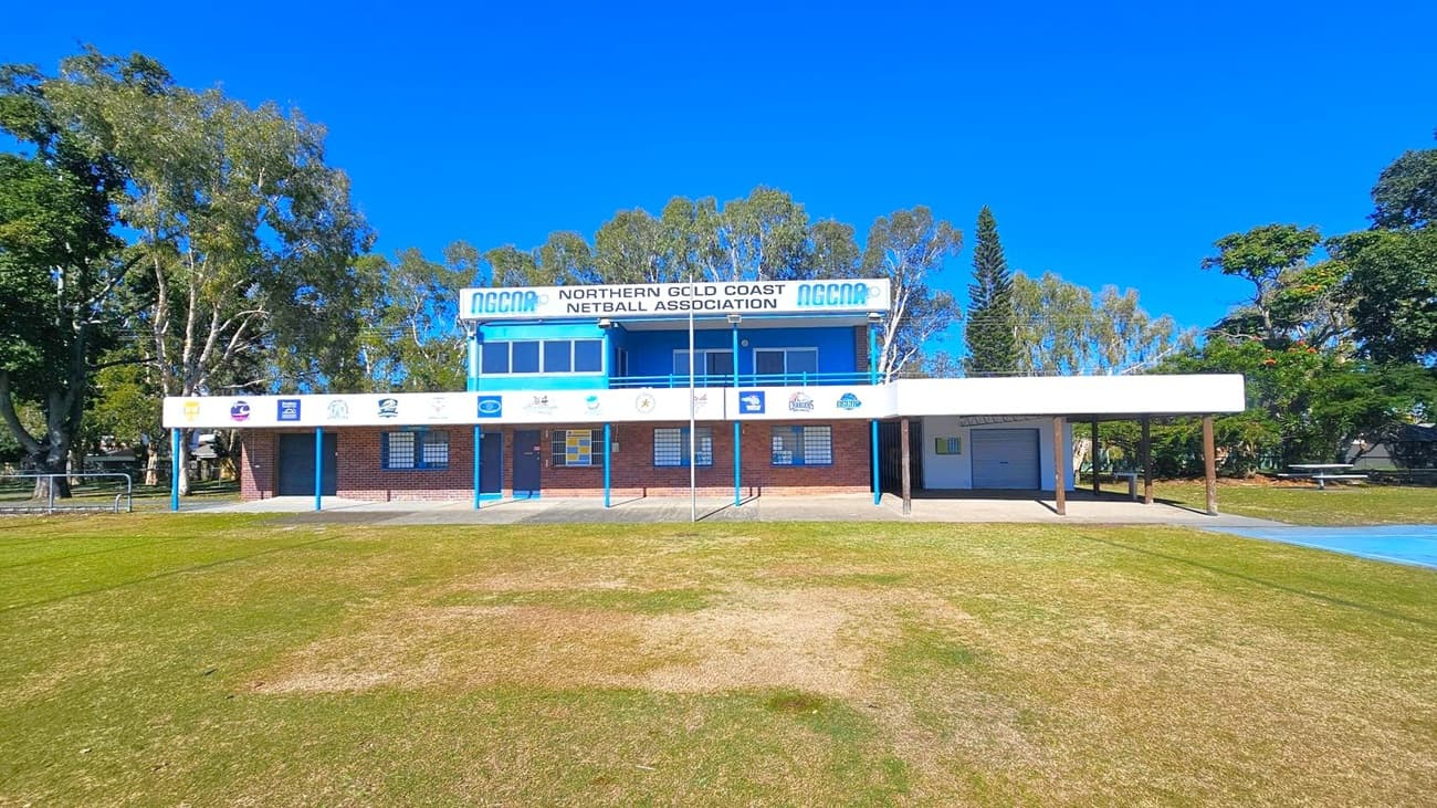 A two-storey building with a sign reading "Northern Gold Coast Netball Association" on the upper level. The structure has a brick facade with blue accents and several windows. The lower level features a series of doors and windows, with various logos displayed above. In the foreground, there is a grassy area with patches of dry grass. Trees surround the building, providing a natural backdrop.