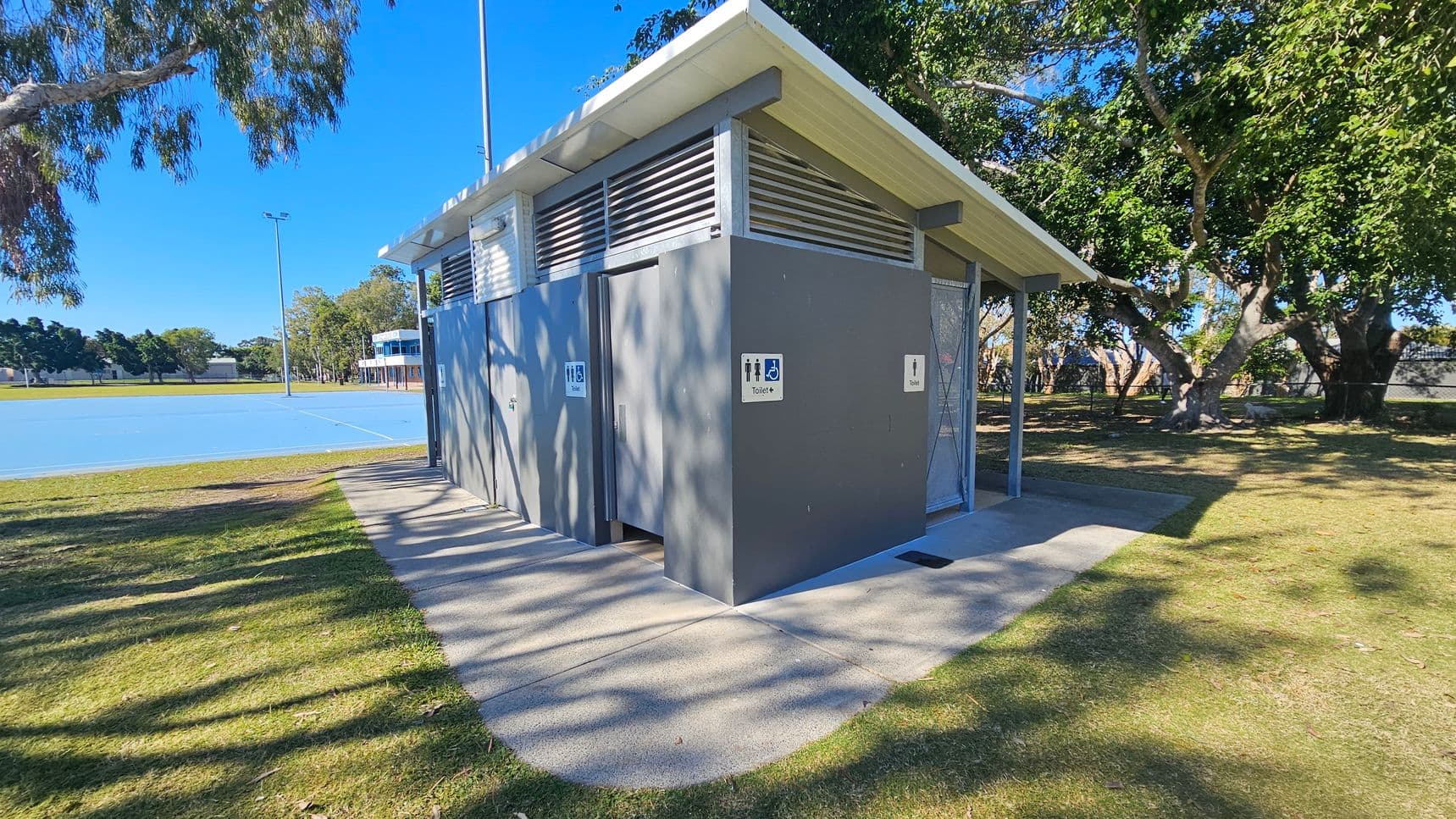 A public toilet block with a slanted roof is situated on a grassy area. The structure is grey with signs indicating male, female, and accessible facilities. To the left, there is a blue sports court with a tall light pole. In the background, trees provide shade, and a building is visible near the court. The ground is a mix of grass and concrete paths, and the area is well-lit by natural light.