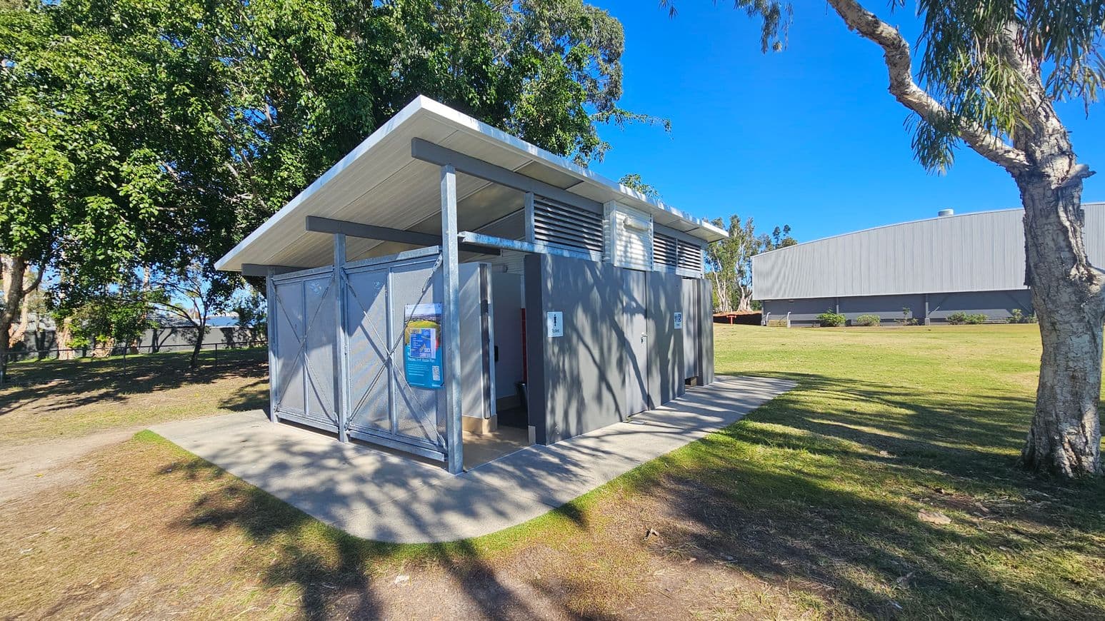 A small public restroom building with a slanted roof is situated on a grassy area. On the left, there are large trees providing shade. The building has metal doors and a sign indicating male facilities. A poster is displayed on the exterior wall. To the right, a large grey industrial building is visible in the background. The ground is a mix of grass and concrete paths, and a tree trunk is in the foreground on the right.