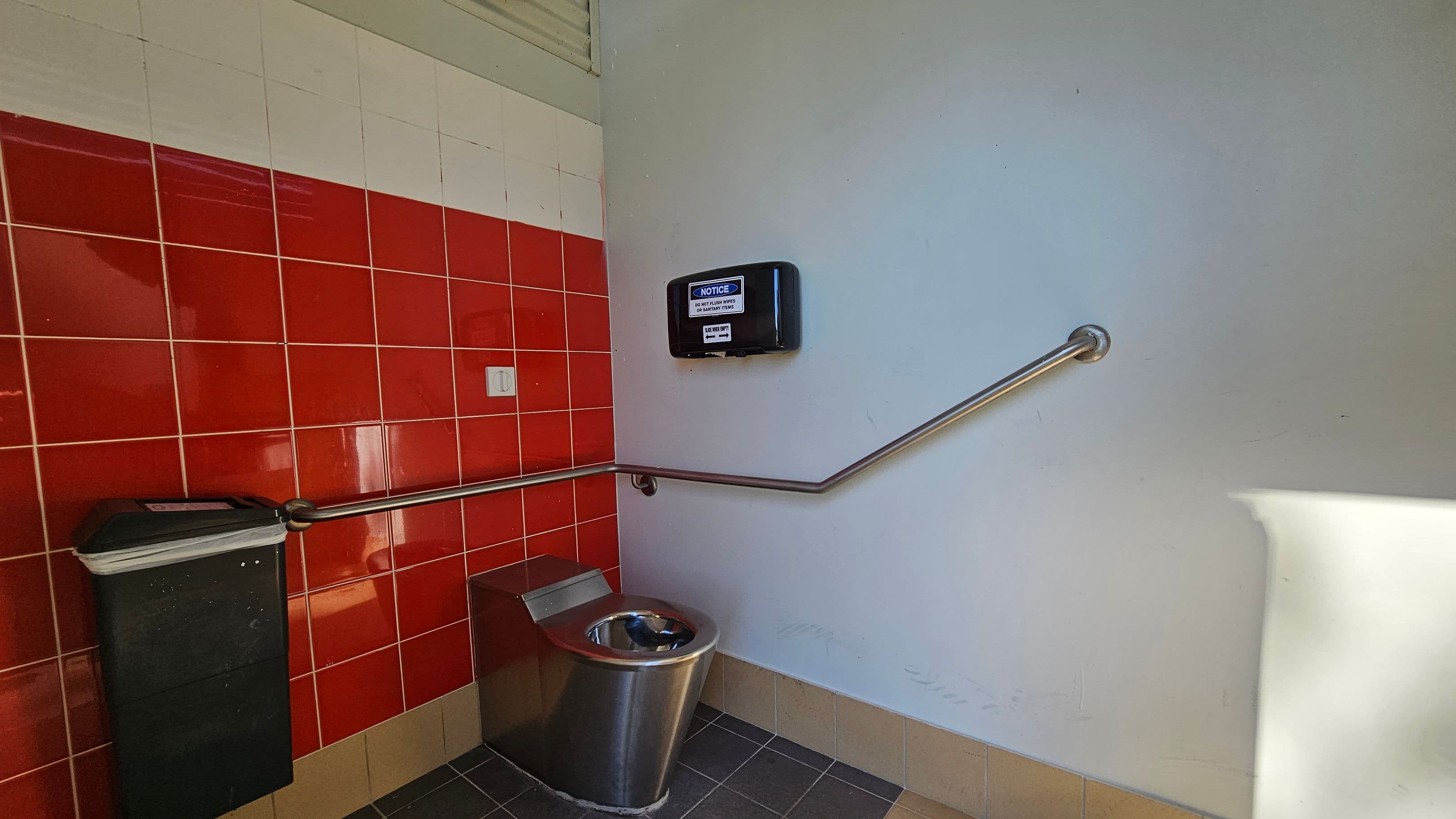 A public restroom with red tiled walls on the left and a light grey wall on the right. A stainless steel toilet is positioned in the corner, with a metal handrail running along the walls. Above the toilet, a black dispenser with a notice is mounted on the grey wall. A black waste bin is attached to the red tiled wall. The floor is covered with dark tiles, and the ceiling is not visible.