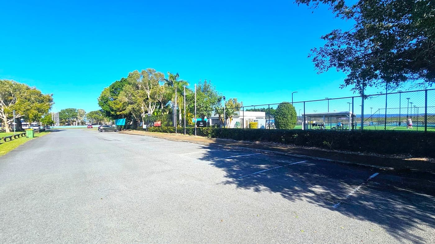A parking area with a few cars is on the left, bordered by a line of trees and a grassy verge with a green bin. On the right, a chain-link fence encloses tennis courts, where people are playing. A small shelter with benches is visible inside the fenced area. The background features more trees and a clear blue sky. Shadows from the trees fall across the parking area.