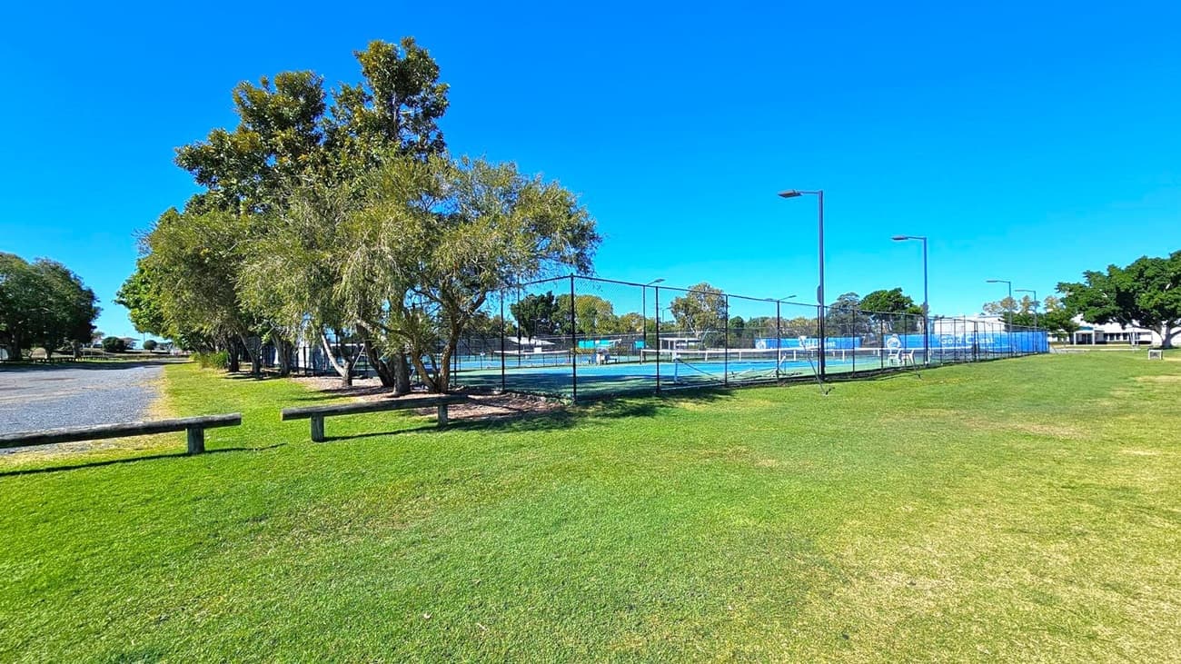 A grassy area with a tennis court enclosed by a chain-link fence on the right. Several tall trees line the left side, providing shade over a gravel path. The tennis court has blue surfaces and is equipped with floodlights. In the background, there are more trees and a white building. The sky is clear and blue.