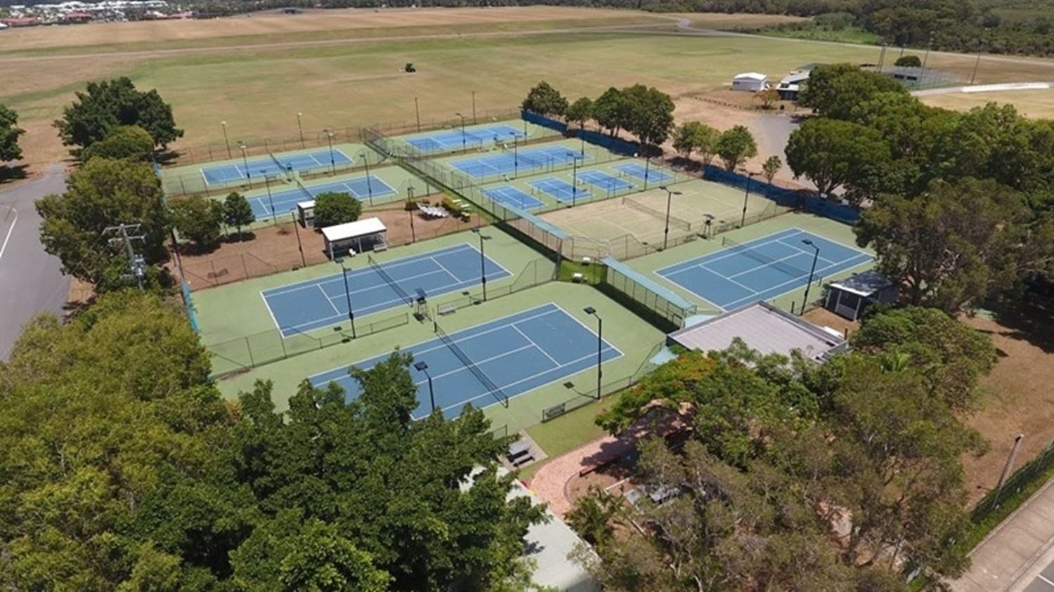 Aerial view of a tennis facility with multiple blue courts surrounded by green fencing. The courts are arranged in rows, with some covered by small shelters. Trees border the area, providing shade and separation from adjacent fields. A few small buildings and structures are visible near the courts. The surrounding landscape includes open fields and a few scattered trees, with a road visible on the left side.