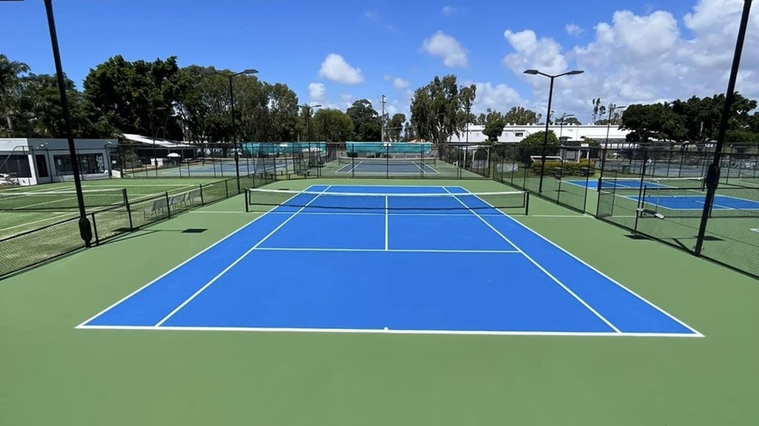 A tennis court with a bright blue playing surface and green surrounding area. The court is enclosed by a black chain-link fence. To the left, there are additional courts and a small building with a shaded area. On the right, more courts are visible. In the background, there are trees and a white building. The sky is partly cloudy, and there are tall light poles around the courts.