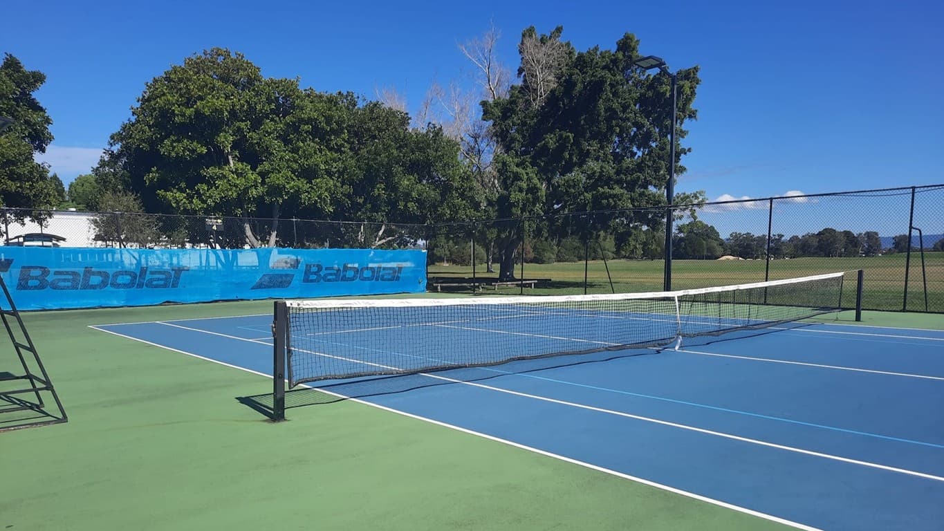 A tennis court with a blue playing surface and green outer area. A white net stretches across the middle. On the left, a metal umpire's chair is visible. In the background, there are trees and a blue banner with the word "Babolat" on a chain-link fence. Beyond the fence, a grassy field extends into the distance.
