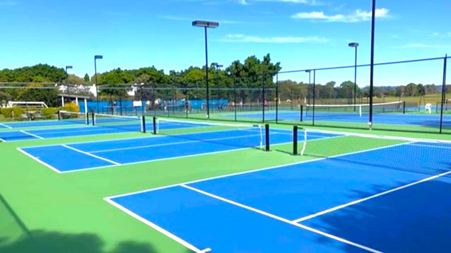 Multiple tennis courts with blue playing surfaces and green borders. The courts are separated by black nets and surrounded by tall black fencing. Several tall light poles are positioned around the courts. In the background, there are trees and a few buildings. The ground is flat, and the area is well-maintained.