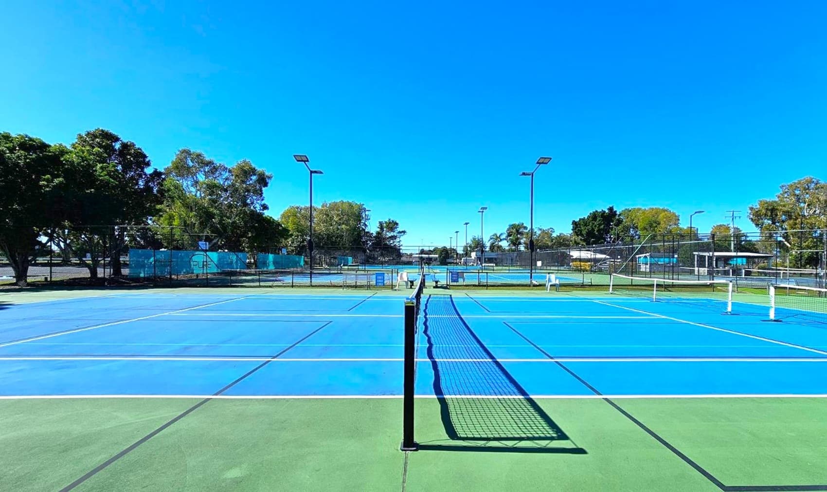 A tennis court with a blue playing surface and green surrounding area. A net runs across the centre. On the left and right, there are tall floodlights and white chairs. In the background, trees and a fence line the perimeter, with additional courts visible beyond. The sky is clear and blue.