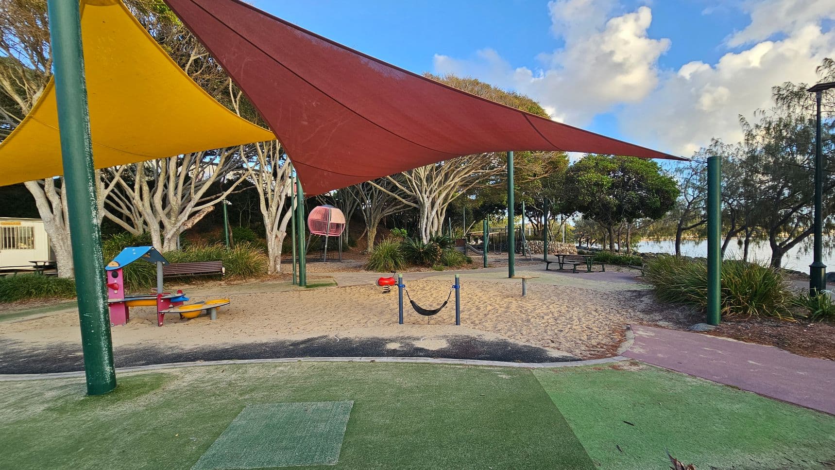 A playground with colourful shade sails, including yellow and red, supported by green poles. On the left, there's a small play structure with a blue roof and a bench nearby. In the centre, a red spring rider and a small hammock are set in a sandy area. To the right, a picnic table is visible under trees. The background features trees and a glimpse of water. The ground is a mix of sand and green artificial turf.