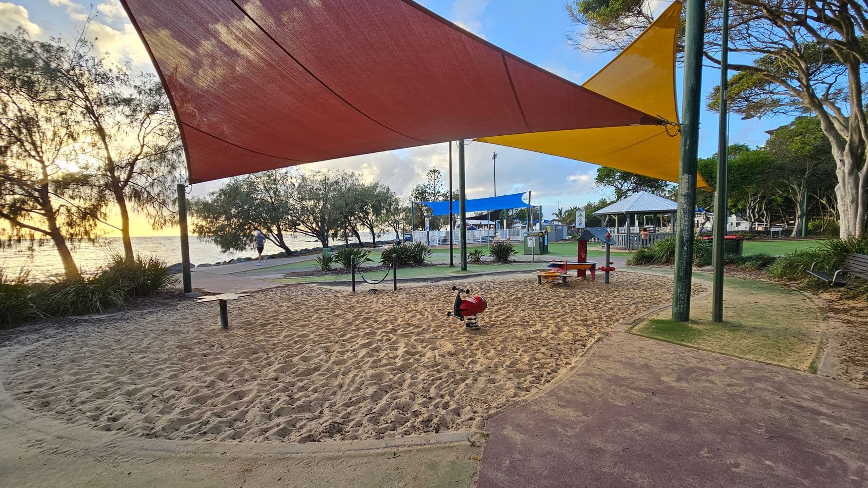 A playground with a sandy area in the centre, featuring a red spring rider. Overhead, there are large triangular shade sails in red and yellow. To the right, a bench and trees are visible. In the background, a person is jogging along a path near the water, with more trees and a gazebo further back. The ground is a mix of sand and coloured pathways.