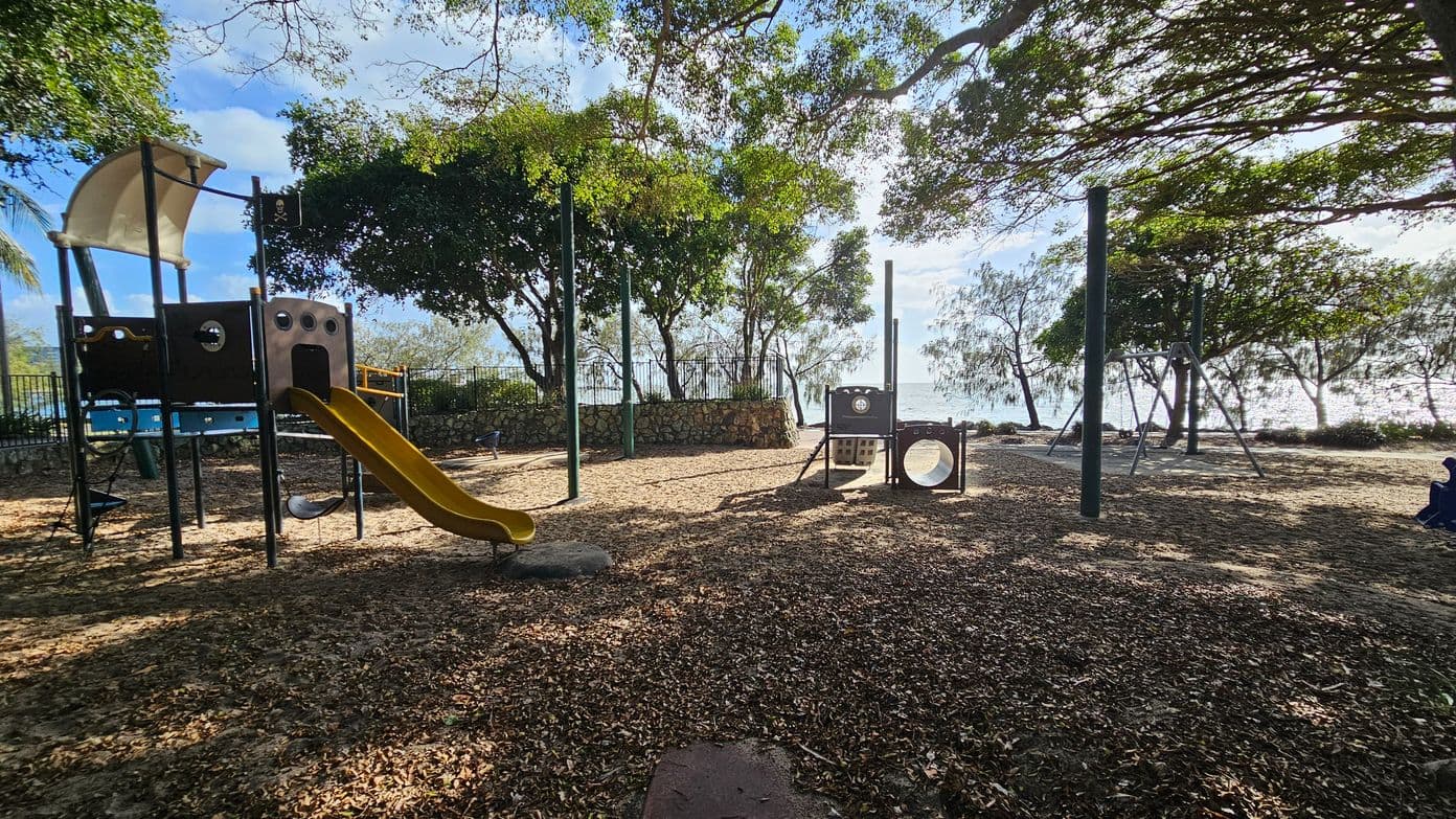A playground with a pirate ship-themed play structure on the left, featuring a yellow slide and a sail. In the middle, there's a small climbing structure with a tunnel. The ground is covered with dry leaves and dirt. Trees with dense foliage provide shade, and a metal fence runs along the background, with a view of the sea beyond. A swing set is visible on the right, and a person is seated on the ground in the far right corner.