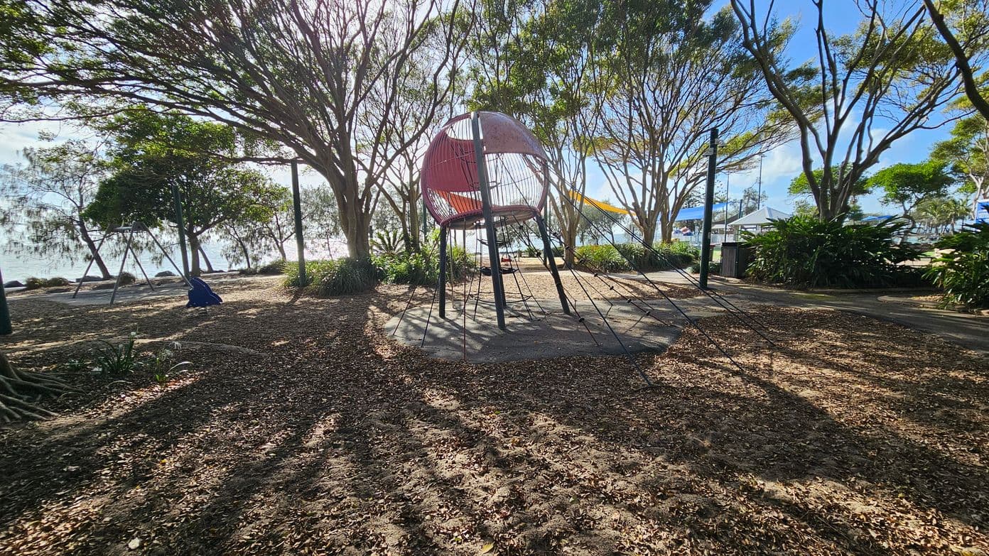 A playground with a spherical climbing structure in the centre, supported by metal poles and surrounded by ropes. To the left, there is a swing set with a blue seat. The ground is covered with dry leaves and dirt. Tall trees with sparse foliage are scattered throughout, casting shadows on the ground. In the background, there is a glimpse of water and more trees, with a clear sky above.