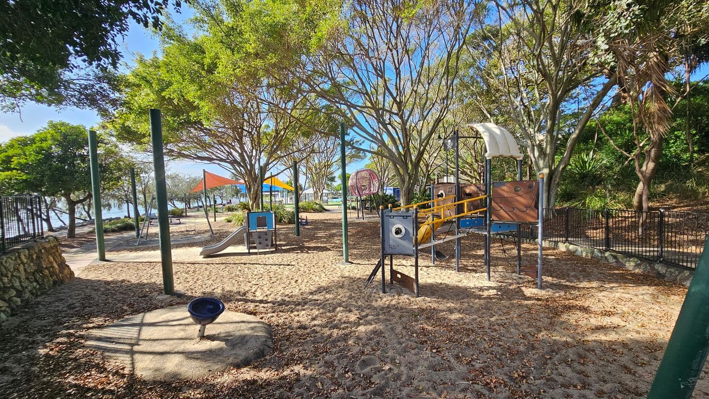 Playground with various equipment surrounded by trees. On the right, a play structure with a slide and a pirate-themed design. In the centre, a climbing frame and a small slide. To the left, a spinning bowl and a path leading to a picnic area with tables under orange shade sails. The ground is covered with sand and fallen leaves. A black metal fence runs along the right side, and the background features more trees and a glimpse of water.