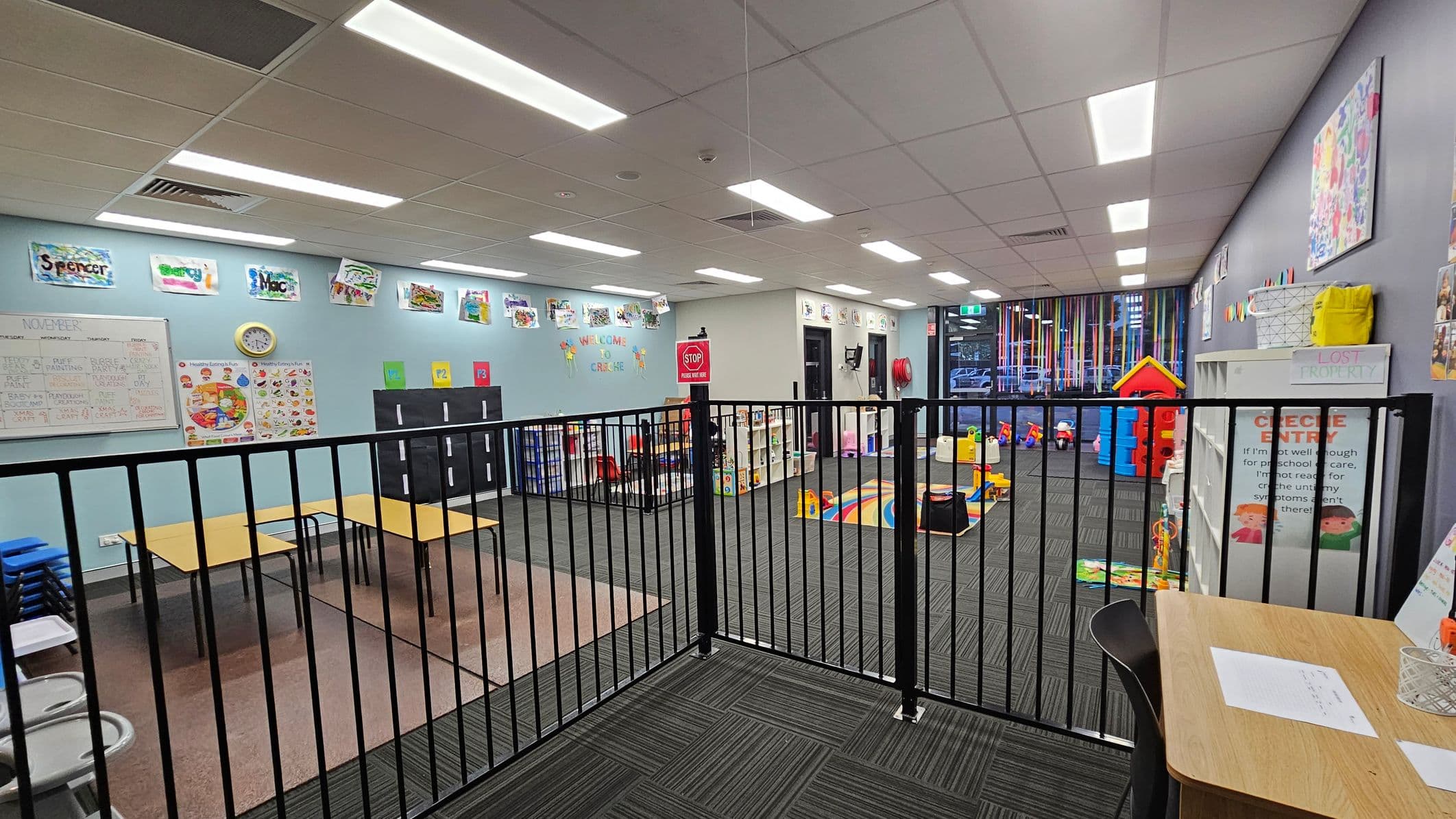 A brightly lit childcare room with a black safety gate in the foreground. On the left, there are tables and chairs, educational posters, and a clock on a light blue wall. The middle area has a colourful play mat and toys, with a "Welcome to Creche" sign above. On the right, shelves hold various items, and a small red playhouse is visible. The ceiling has recessed lighting, and the floor is carpeted in dark grey.