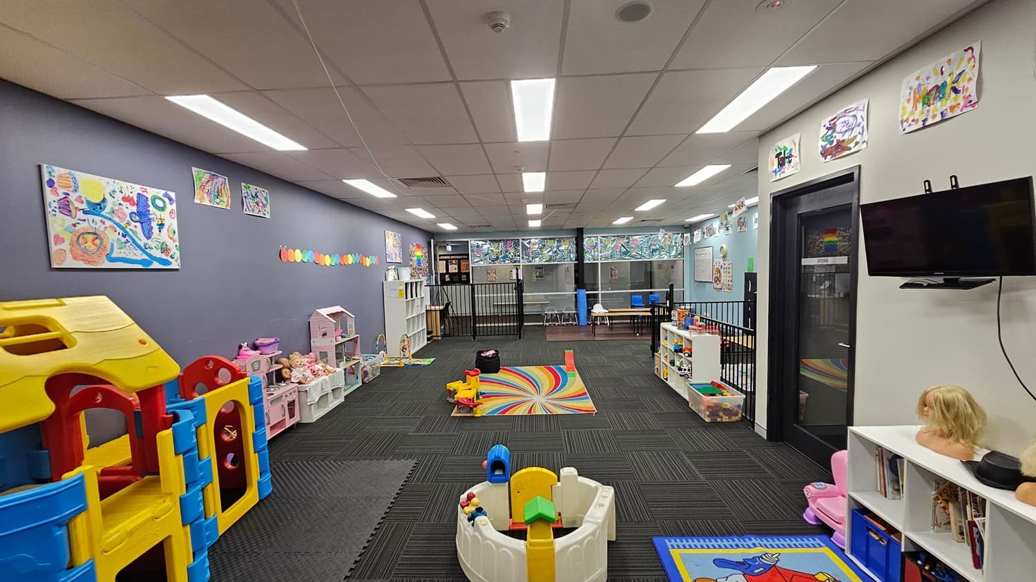 A colourful playroom with a variety of toys and activities. On the left, there's a large plastic play structure in yellow, red, and blue. Along the left wall are shelves with dolls and a dollhouse. The centre features a rainbow-patterned rug and various toys. On the right, a wall-mounted TV is above a door labelled "STORE". The walls are decorated with children's artwork. The floor is covered with dark carpet tiles, and the ceiling has bright fluorescent lights.