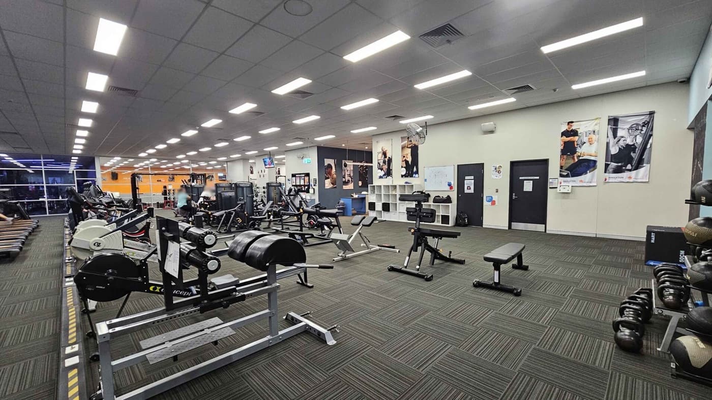 A spacious gym with various exercise equipment. On the left, there are rowing machines and treadmills near large windows. In the centre, weightlifting benches and machines are arranged neatly. The right side features kettlebells and medicine balls on racks. The walls have fitness posters, and there are two doors. The floor is covered with green and grey patterned carpet tiles, and the ceiling has bright fluorescent lights.