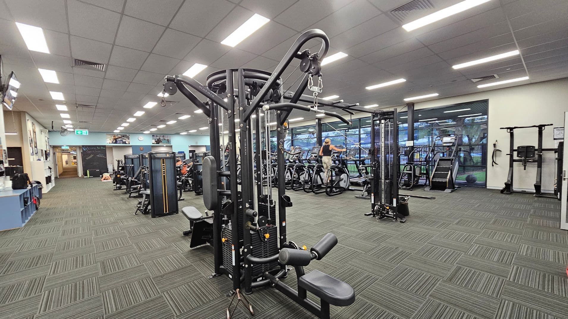 A spacious gym with various exercise machines. In the centre, there's a large multi-functional weight machine. To the right, there are treadmills and exercise bikes near large windows. The left side has a reception area with a counter and a chalkboard wall. The floor is covered with green and grey patterned carpet tiles, and the ceiling has bright fluorescent lights.