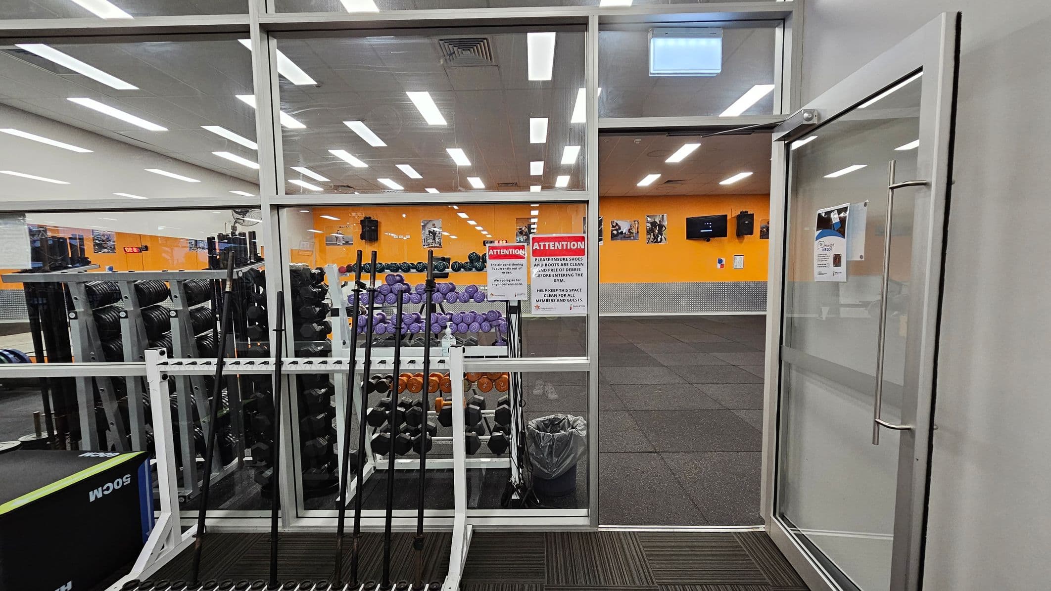 A gym interior with a glass wall and door on the right. Inside, there are racks of dumbbells and weightlifting bars. The walls are orange with several posters and a TV screen. The floor is covered with black rubber mats. Bright ceiling lights illuminate the space. Signs on the glass wall provide instructions for gym users. A trash bin is visible near the weights.