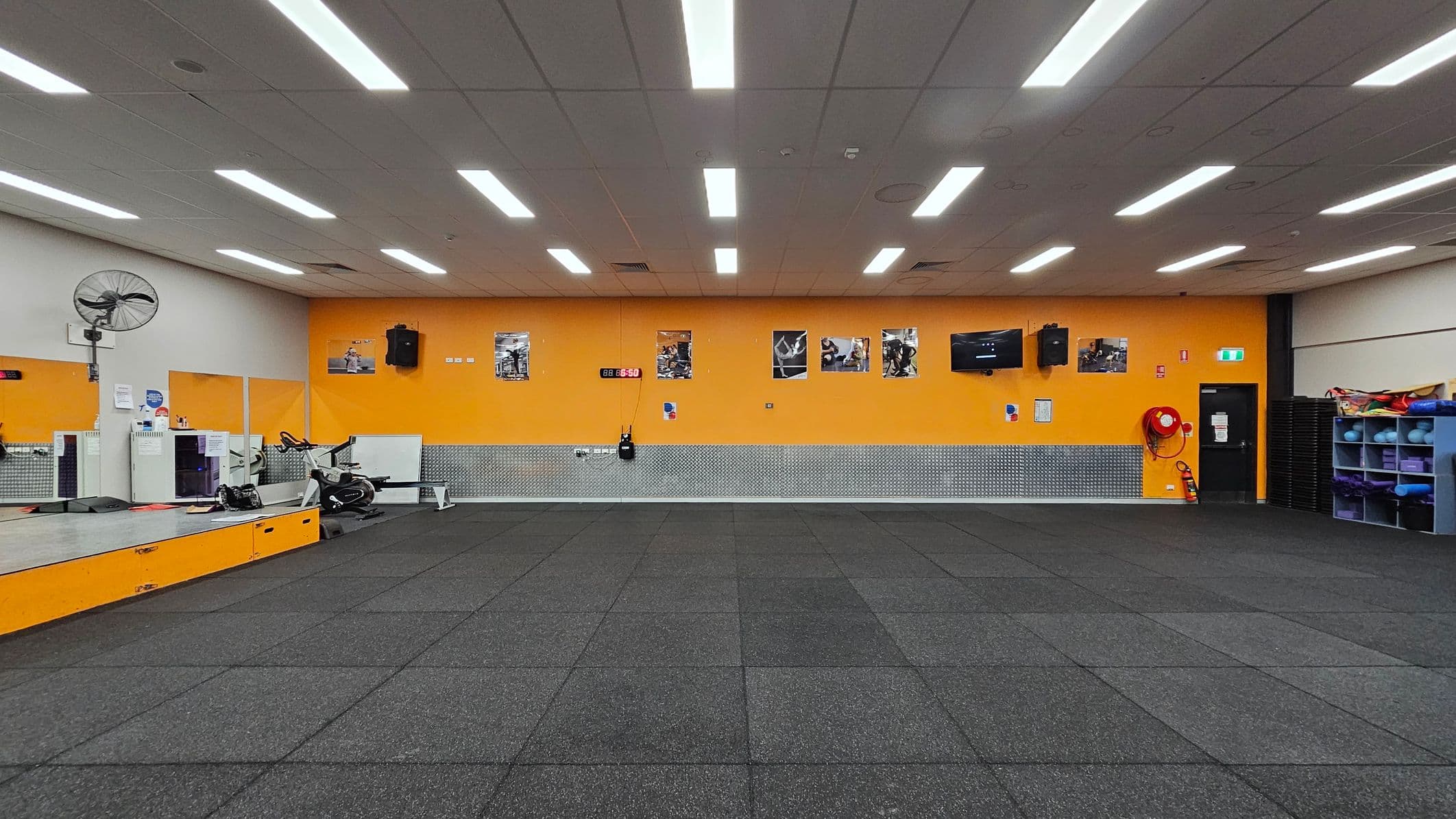 A gym room with a black rubber-tiled floor and a white ceiling with fluorescent lights. The back wall is orange with several fitness posters and a digital clock. On the left, there's a raised platform with a fan, exercise bike, and some equipment. On the right, a black door, fire extinguisher, and shelves with exercise mats and balls. The wall has a metallic panel running along its lower half.