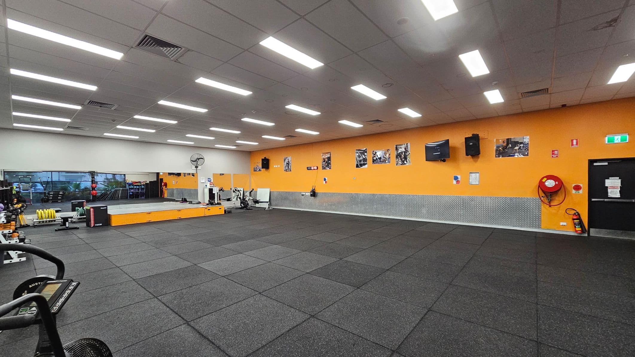 A spacious gym with a black rubber-tiled floor and a white ceiling with fluorescent lights. The left side features a large mirror and various gym equipment, including weights and exercise machines. The back wall is orange, adorned with several posters, a TV, and a speaker. A fire hose reel and an emergency exit sign are visible on the right. A raised platform is positioned near the mirror, and a fan is mounted on the wall.