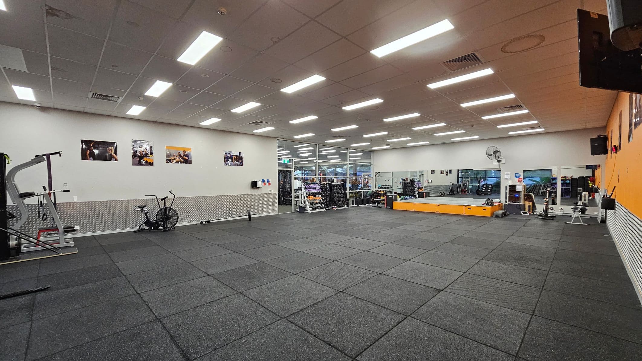 A spacious gym with a grey rubber floor and a white ceiling with fluorescent lights. On the left, there's exercise equipment including a stationary bike and a weight machine. The back wall features several framed photos and a pegboard with hanging items. The right side has a large mirror and a raised platform with more equipment. A fan is mounted on the wall, and a glass partition separates the gym from another area.
