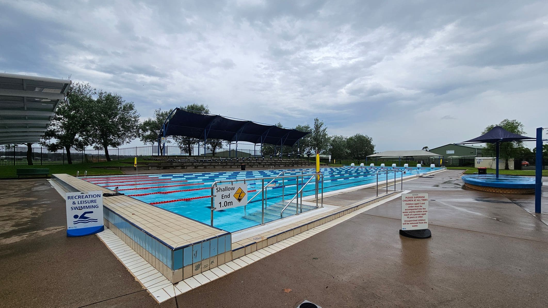 An outdoor swimming pool area with a wet concrete surface. On the left, there's a grassy area with a palm tree and a white pergola. The pool is in the centre, surrounded by a low fence, with blue and white lane dividers. A sign indicates a depth of 1.0m. On the right, there are blue shade structures and a small building in the background. Trees and a fence line the perimeter.