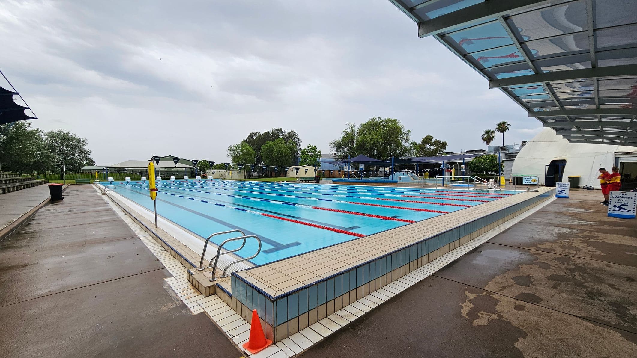An outdoor swimming pool with multiple lanes, marked by red and blue lane dividers. On the left, there is a wet concrete walkway with a black bin and a lifebuoy. On the right, a covered area with a transparent roof and two people in red uniforms standing near signs that read "Lap Swimming." In the background, there are trees, a fence, and some buildings. The pool's edge is tiled, and a small orange cone is placed at the corner.