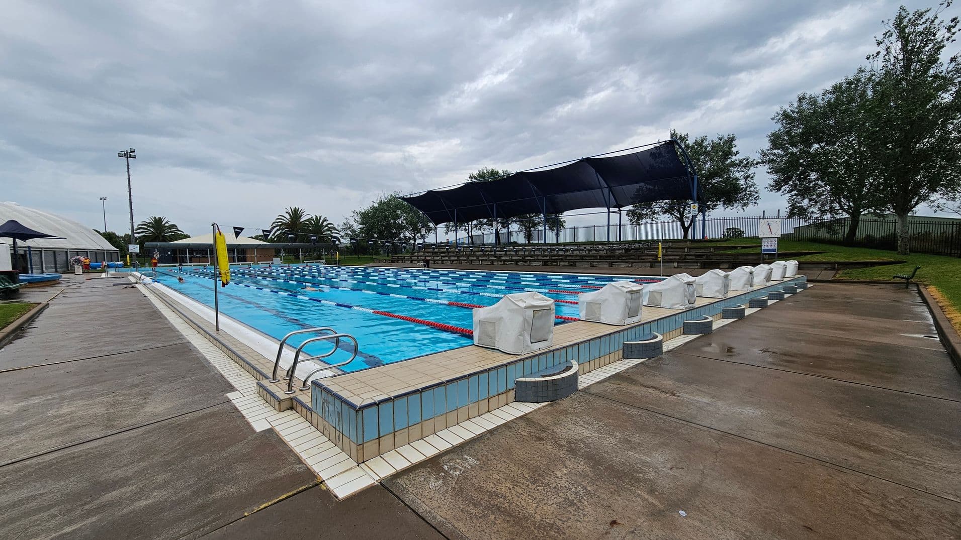 Outdoor swimming pool with multiple lanes, surrounded by a wet concrete deck. On the right, there are covered starting blocks and a grassy area with trees and a fence. On the left, a pathway leads to a building with a dome-shaped roof and palm trees nearby. A row of shaded seating is visible in the background. The pool has blue and white lane dividers, and a metal ladder is at the pool's edge. The sky is overcast.