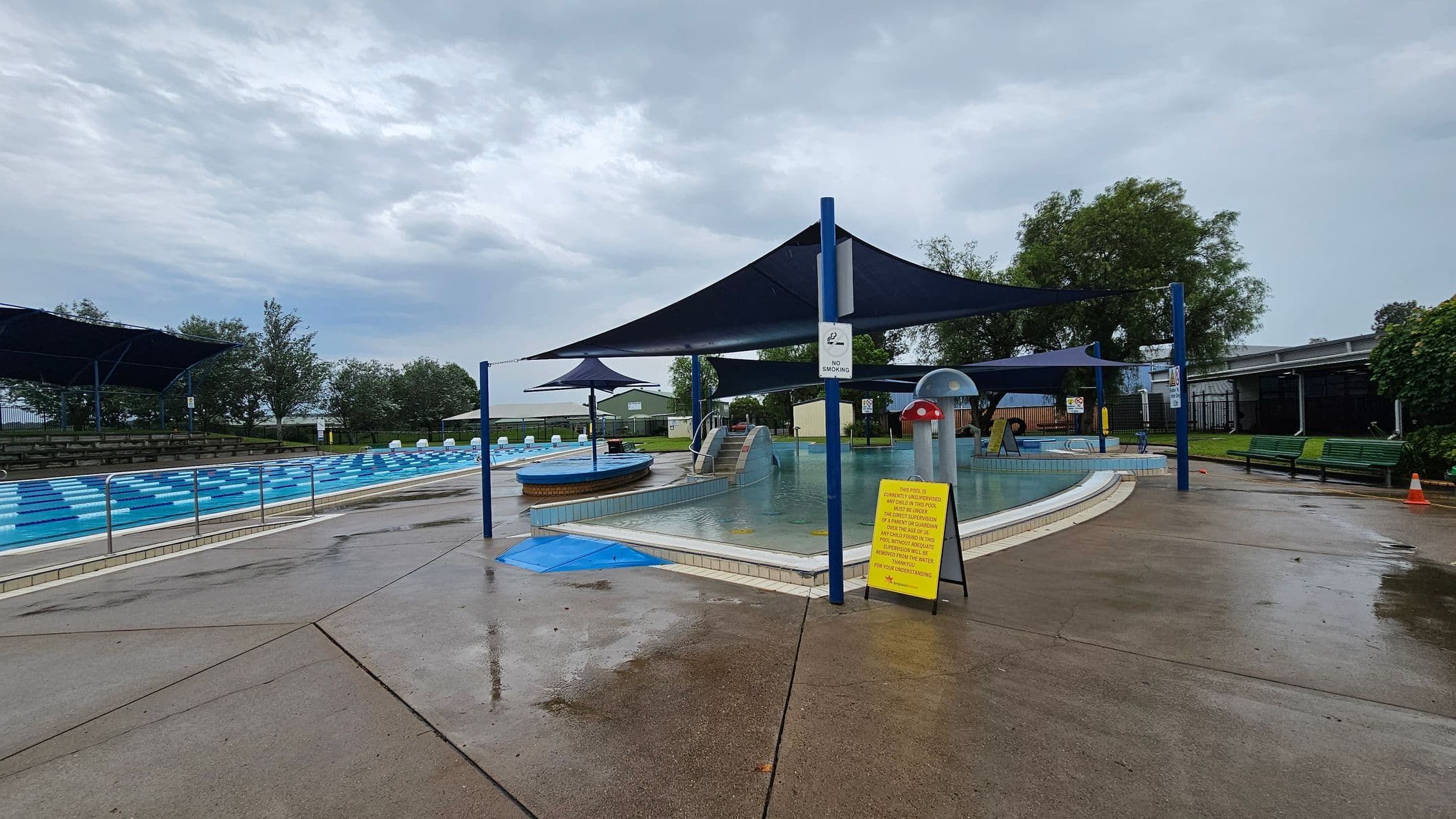 A swimming pool area with a wet concrete floor. On the left, there is a large pool with blue and white lane dividers, and a shaded seating area with trees in the background. In the centre, a smaller pool features a slide and a mushroom-shaped water feature, covered by blue shade sails. A yellow sign with pool rules is visible. On the right, there are green benches and more trees, with a building partially visible in the background.