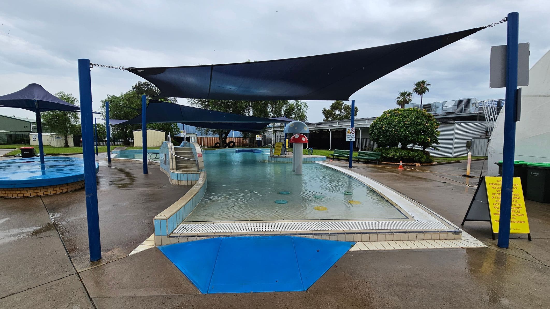 A children's splash pool area with a shallow water feature in the centre, surrounded by a tiled edge. A mushroom-shaped water fountain is visible in the middle. Blue shade sails are suspended above, supported by blue poles. To the right, a yellow sign is placed on the wet concrete ground. In the background, there are trees, a building with metal siding, and a grassy area with a bush. The sky is overcast.