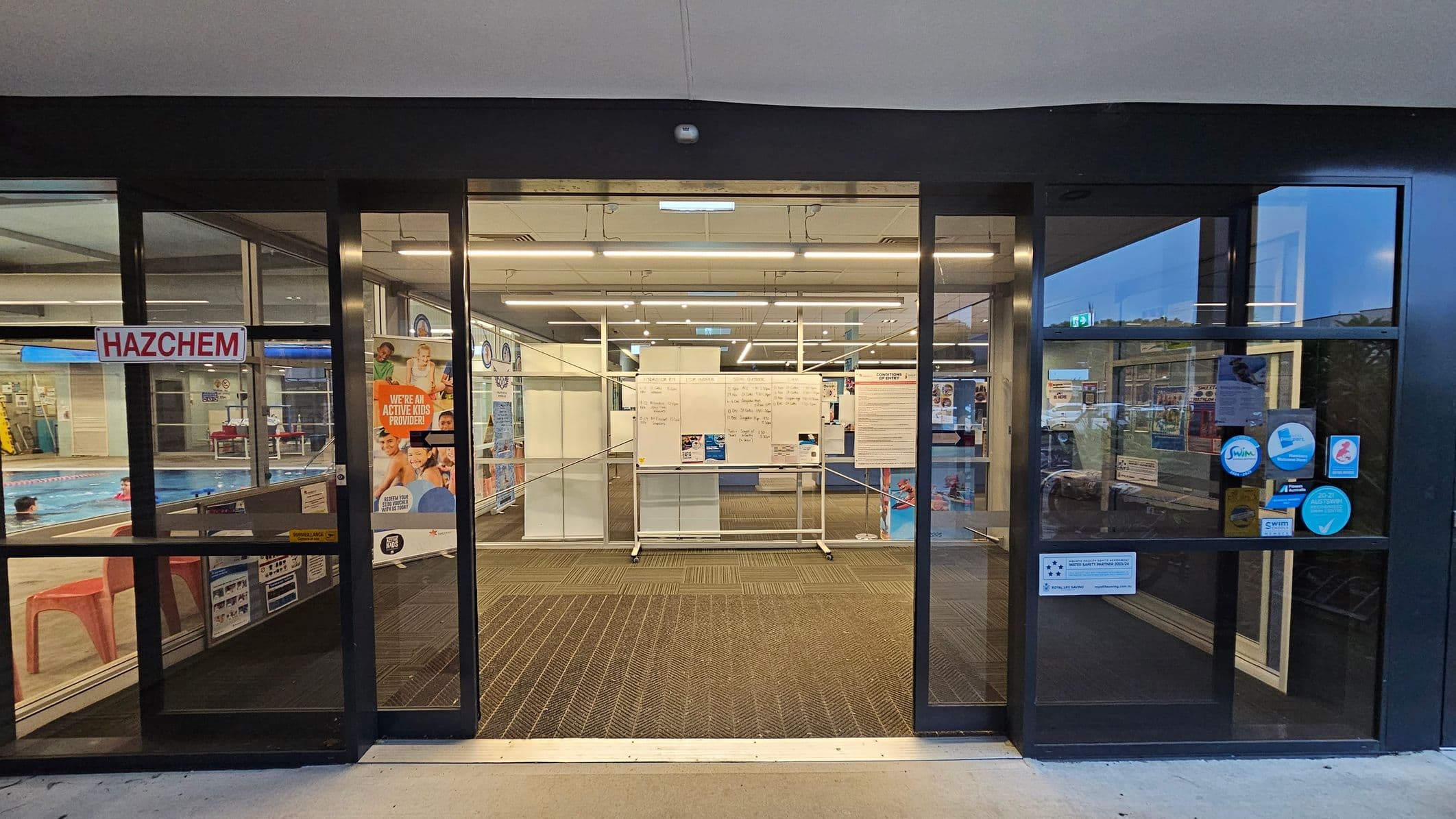 Glass entrance doors to a swimming pool facility. On the left, a "HAZCHEM" sign is visible, with a glimpse of the pool and swimmers inside. On the right, several stickers and notices are displayed on the glass. In the centre, a whiteboard with information is positioned behind the doors. The floor is carpeted with a patterned mat, and the ceiling has fluorescent lighting. Posters and promotional materials are visible inside.