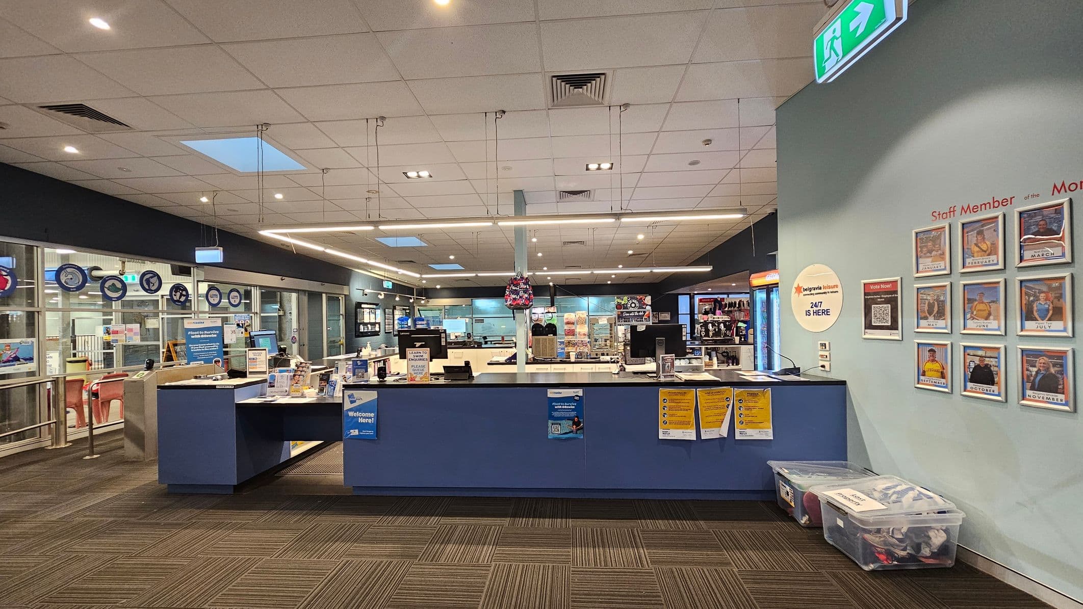 A reception area with a blue counter in the centre, displaying various informational materials and a small backpack hanging above. Behind the counter are computer monitors and a well-lit interior with shelves and displays. On the right wall, there are framed photos labelled with months, titled "Staff Member of the Month." Below, clear plastic bins are placed on the floor. The ceiling is fitted with lights and ventilation panels, and the floor has a patterned carpet.