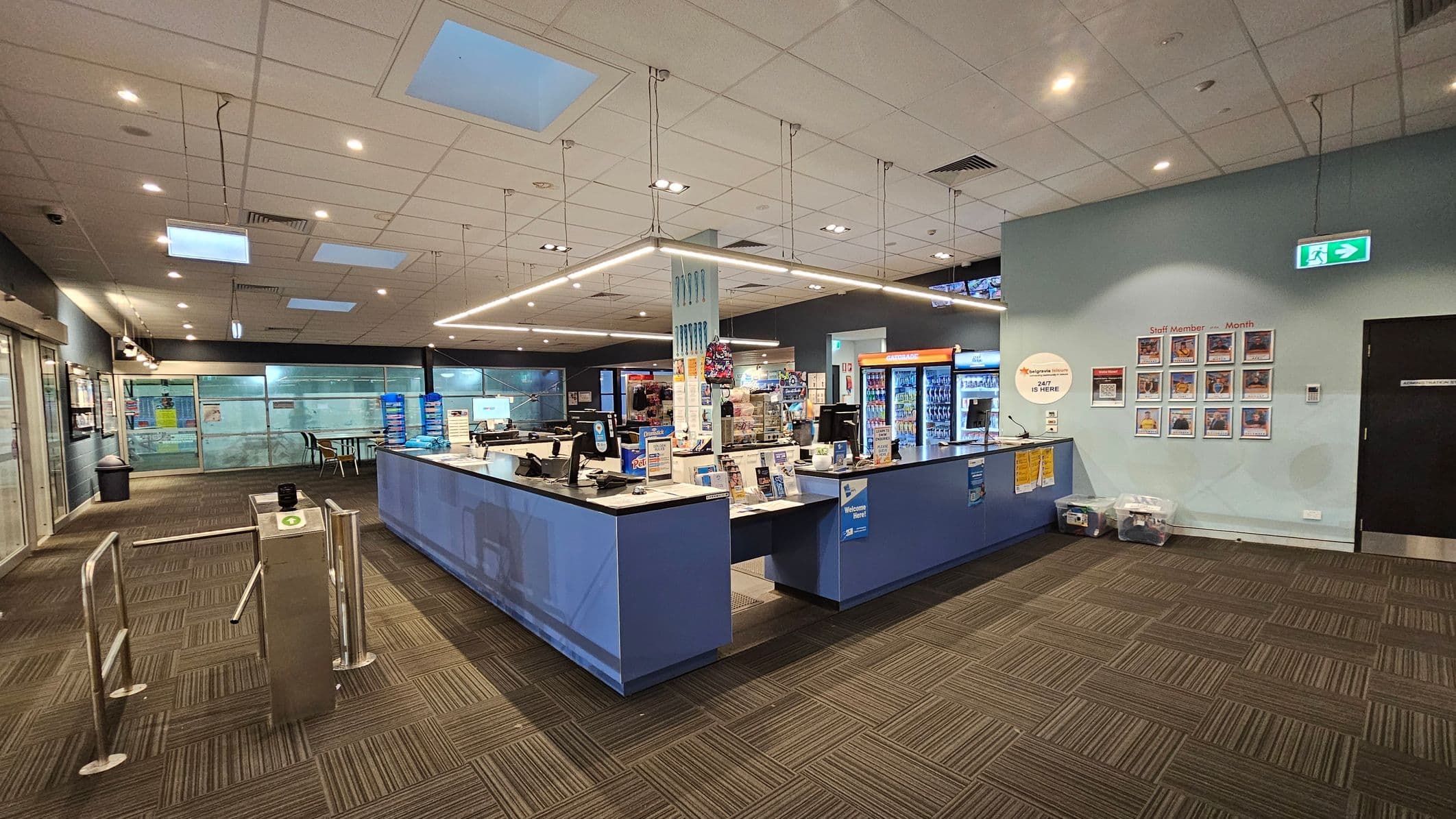 A spacious interior with a reception desk in the centre, featuring computers and promotional materials. To the right, a wall displays "Staff Member of the Month" photos and an emergency exit sign. On the left, a turnstile and glass doors lead to another area. The floor is carpeted with a geometric pattern, and the ceiling has recessed lighting and skylights. Vending machines and seating are visible in the background.
