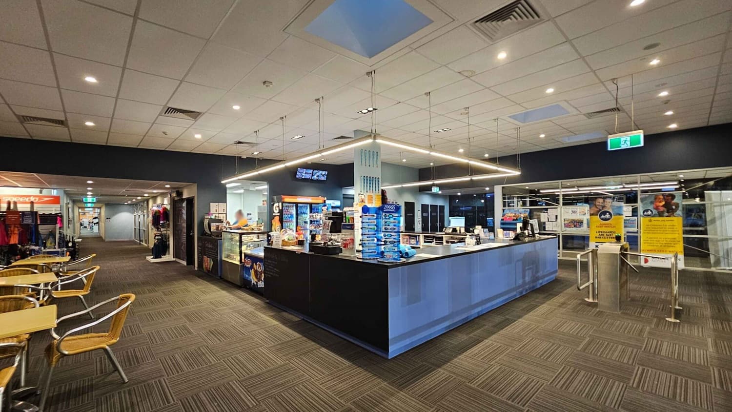 A spacious indoor area with a modern reception counter in the centre, displaying various products and a cash register. To the left, there are tables with wicker chairs and a display of clothing. The floor is carpeted with a geometric pattern. The ceiling is fitted with recessed lighting and a skylight. On the right, there are large glass windows with posters and a turnstile entrance. The background features a corridor leading to other areas.