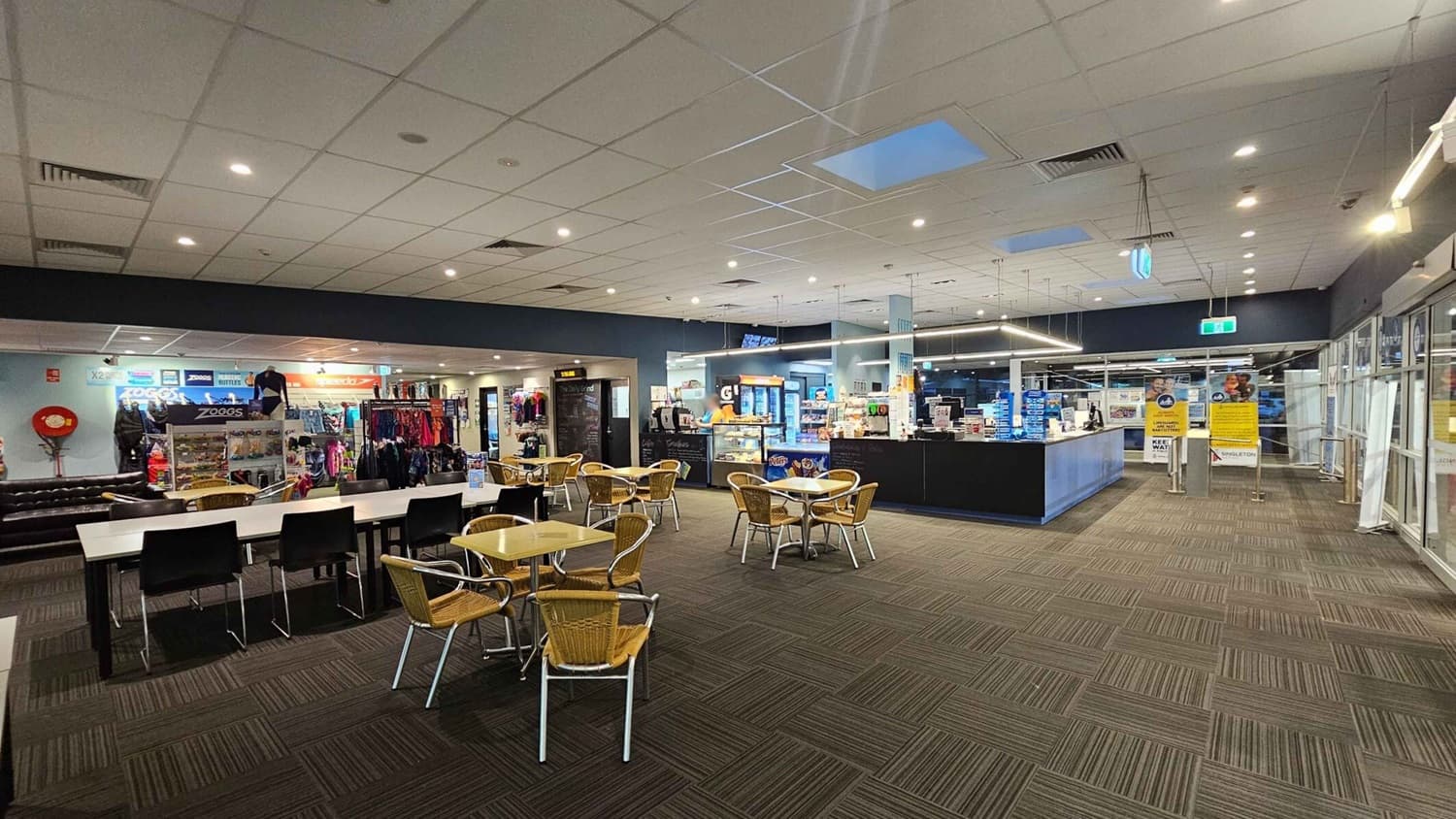 A spacious indoor area with a tiled ceiling and carpeted floor. In the foreground, there are tables with wicker chairs. On the left, a shop displays swimwear and accessories with brands like Zoggs and Speedo. In the centre, a café counter offers snacks and drinks. On the right, large glass doors and windows lead to another area, with signs and notices visible. The lighting is bright and evenly distributed.